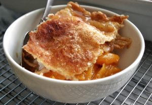 A picture of peach cobbler with a fllaky crust in a white bowl with a spoon.